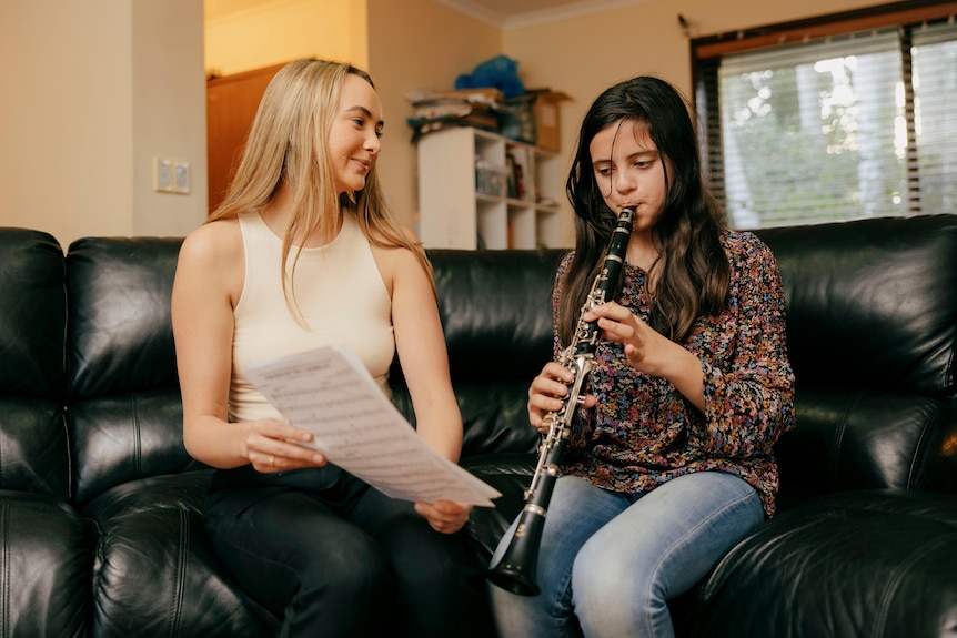 Woman sitting on a black leather couch with a music manuscript helping a young girl practice the clarinet